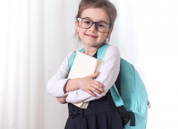 Girl-elementary school student with a backpack and a book with glasses is on a light background .The concept of education and primary school